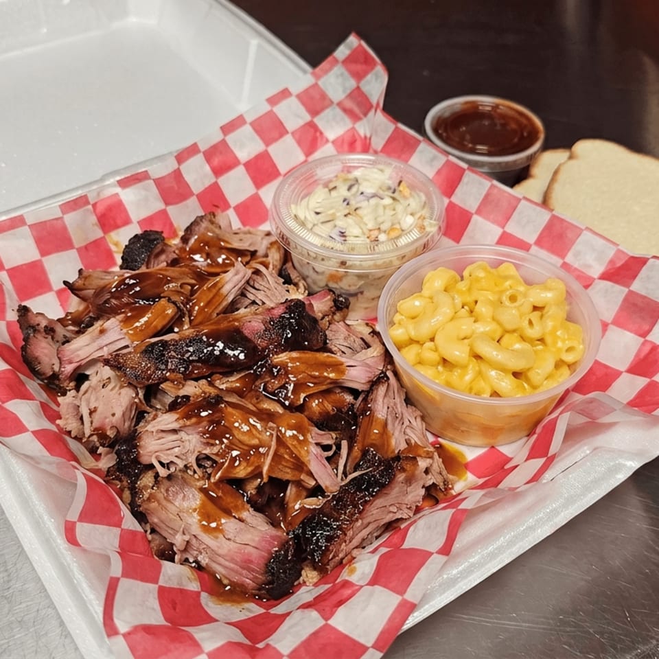 Sliced smoked barbecue meat with sauce, macaroni and cheese, coleslaw, bread, and dipping sauce in a checkered-lined takeout tray.
