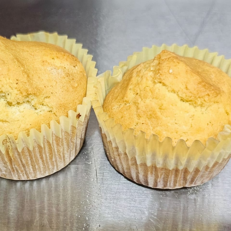 Two golden muffins in paper liners rest on a scratched metal countertop.
